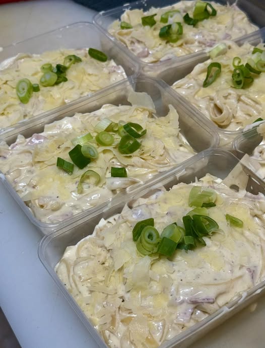 Creamy bacon carbonara pasta with parmesan cheese and fresh spring onion, served in a takeaway container.